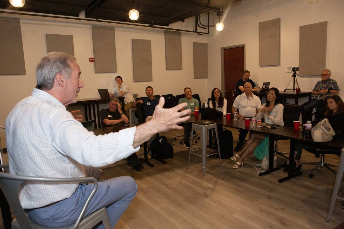 Mike seated, arm extended, talking with a room of about twenty people around tables.
